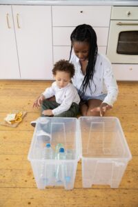 A mother and her child sort plastic bottles into recycling containers on a wooden kitchen floor.
